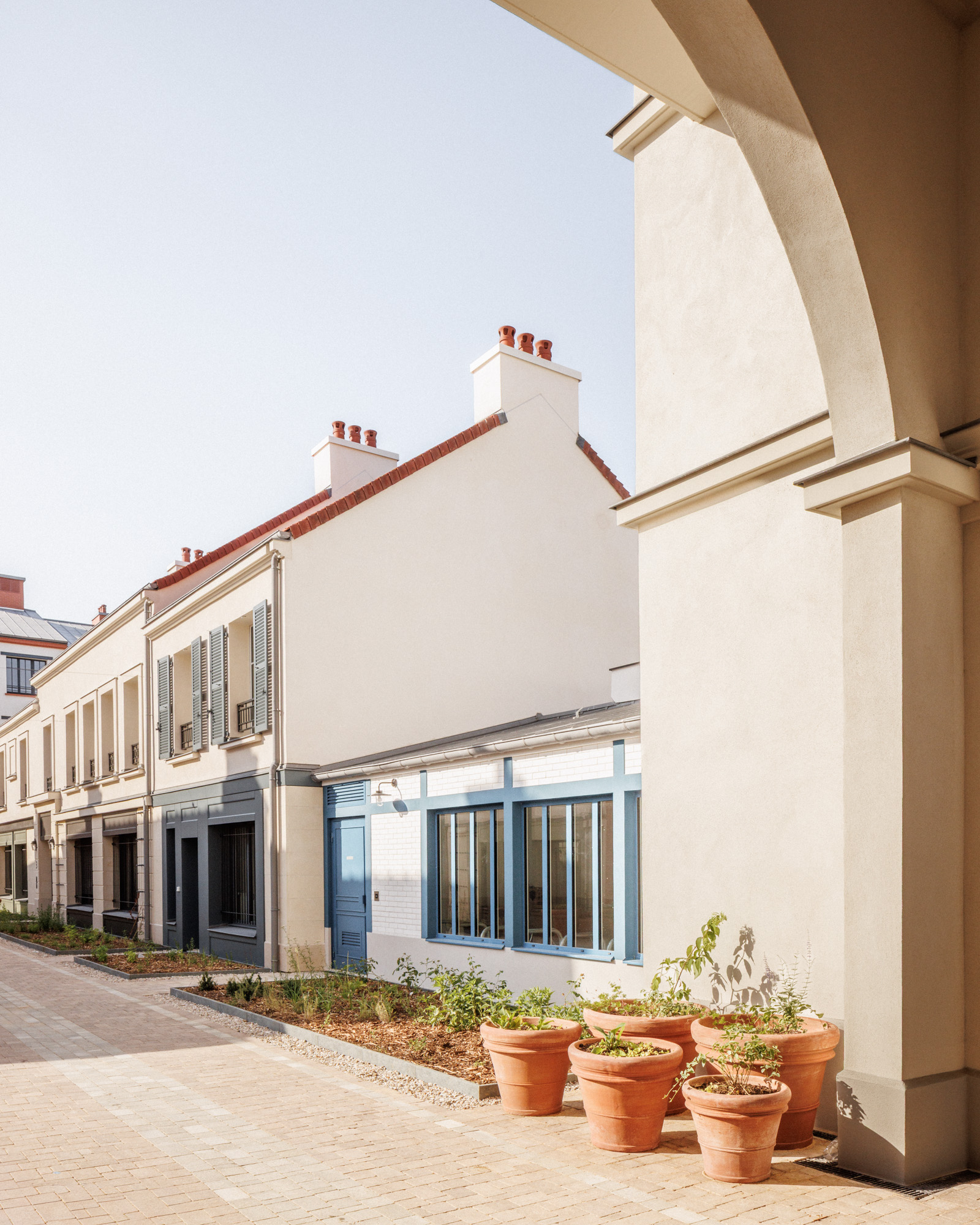 Vue depuis le porche sur le local vélo aux menuiseries bleu ciel et sur des pots en argile de végétation sur le projet de logements sociaux de Serris Magellan par Cussac Architectes et Associés (CA+A)